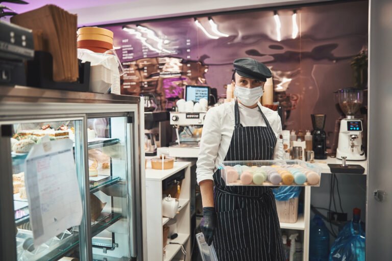 Responsible lady in chef uniform wearing mask and holding a container full of macarons while posing at cozy cafe